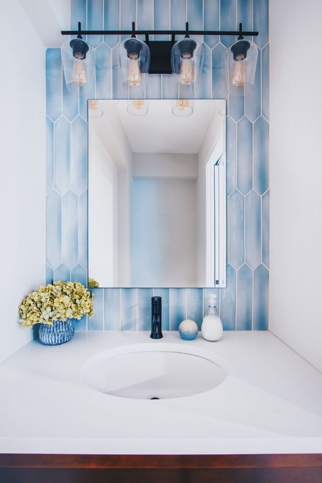 Blue tile behind bathroom mirror with white countertop and dark light fixture